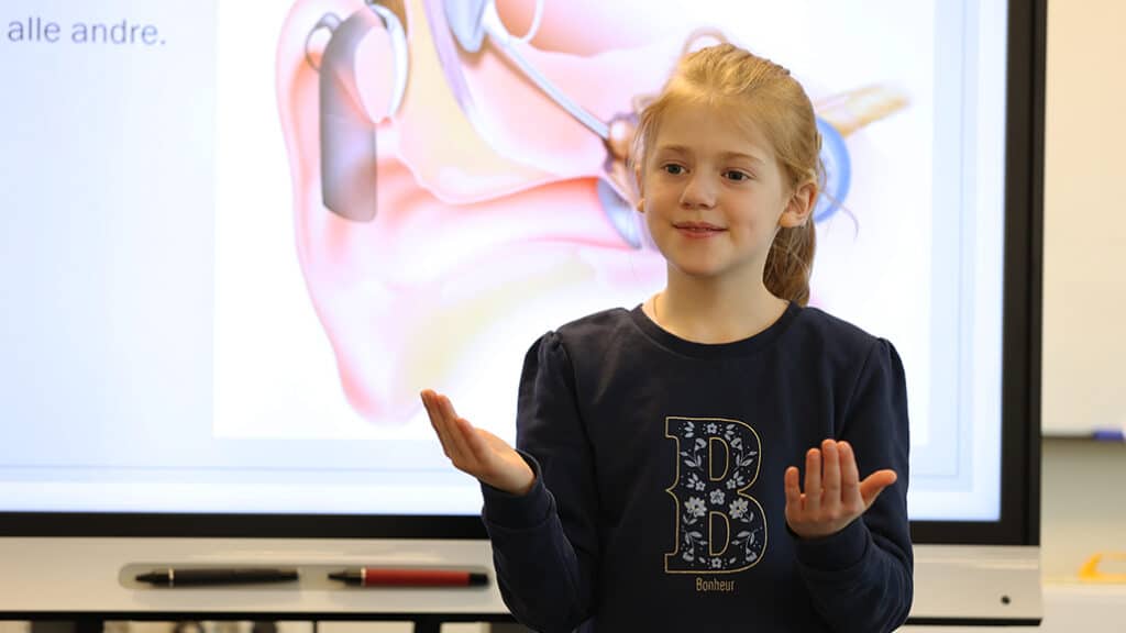 Emma bruker tegnspråk foran tavle med bilde av øre
