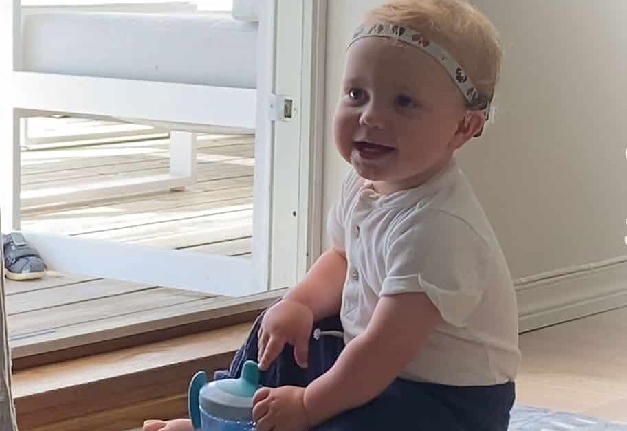 Tidemand (1) med CI sitter på en matte med drikkekopp. Tekst: "Små ører store muligheter. På vei til hørsel, språk og kommunikasjon"