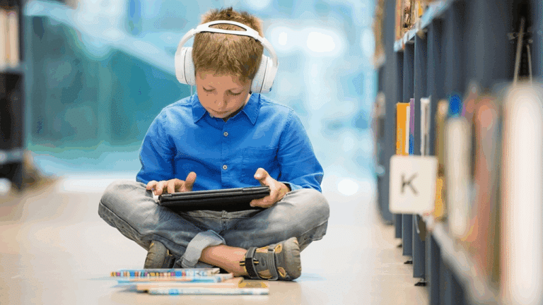 Gutt med hodetelefon og nettbrett sitter ved bokhylle og hører på skolelydbøker