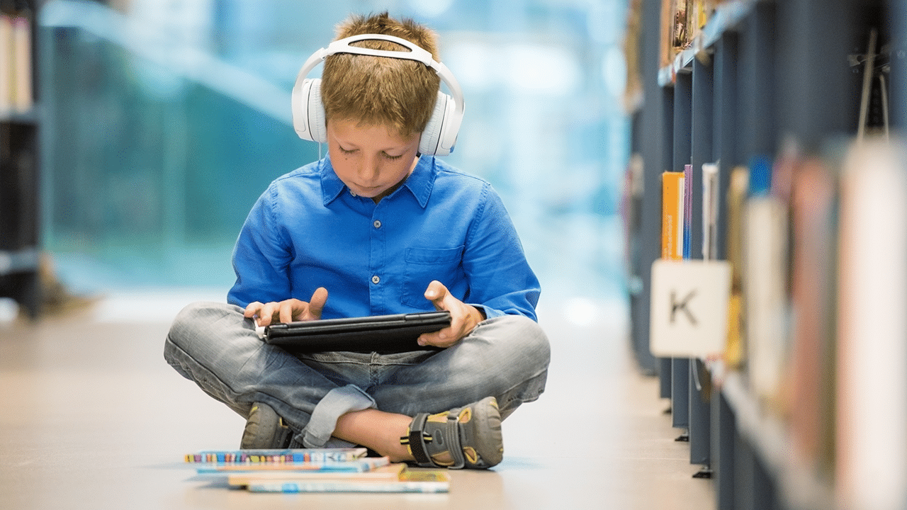 Gutt med hodetelefon og nettbrett sitter ved bokhylle og hører på skolelydbøker