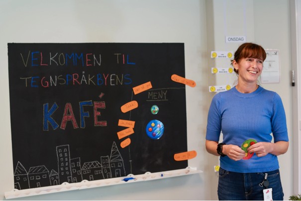 Lærer fra deltidsopplæringen står foran sort tavle der det står "Velkommen til tegnspråkbyens kafe"