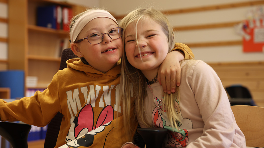 Pernille holder rundt venninne mens de sitter i klasserommet. Begge smiler